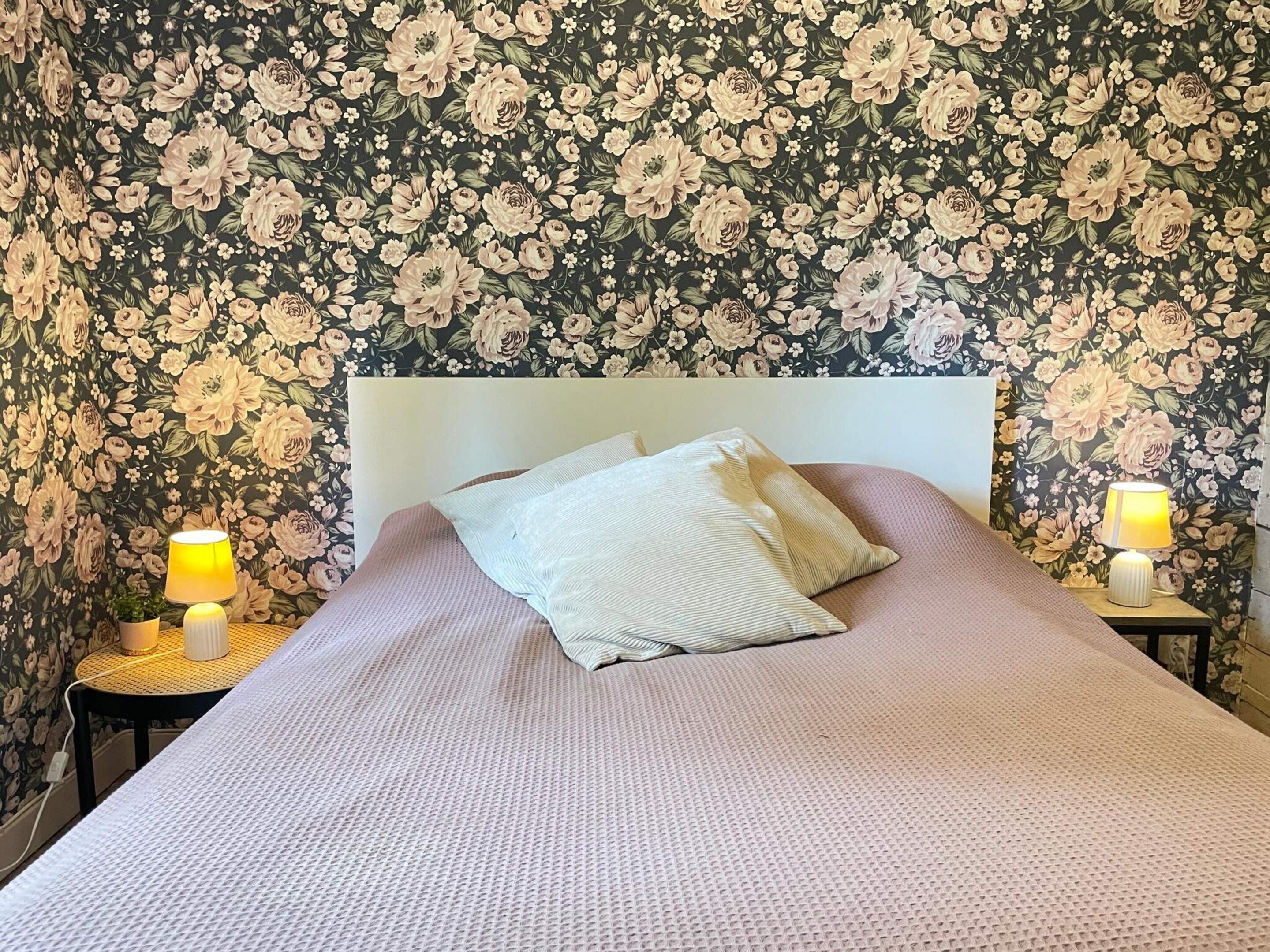 A bedroom with a floral wallpaper, a white headboard, a pink bedspread, two bedside lamps, and a small round table with a lamp.