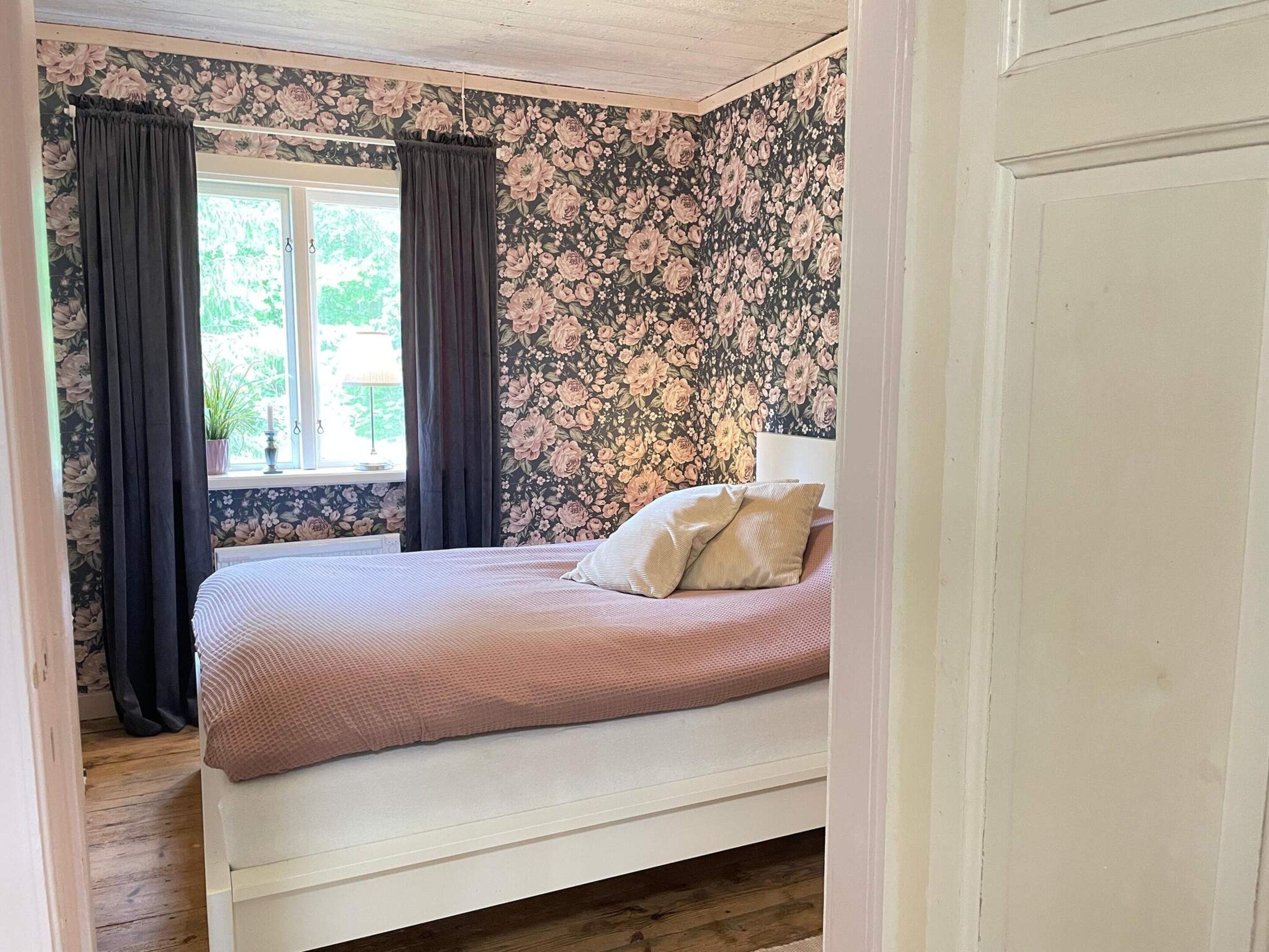 Une chambre à coucher avec un lit, des rideaux, un papier peint à motifs floraux et une fenêtre.