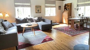 A cozy living room with a sofa, a round coffee table, and a dining table with chairs. There are windows with blinds, a chandelier, and a wooden ceiling. - 4 Person Holiday Home in Arkelstorp (Arkelstorp)