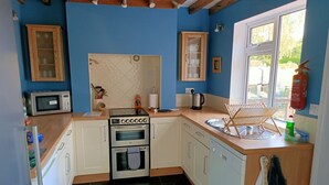 Een kleine keuken met een magnetron, oven en gootsteen. Houten keukenkastjes en een raam zorgen voor natuurlijk licht.