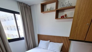 A neatly made bed with a striped blanket and two pillows, a wooden headboard, a small wooden nightstand, and a shelf with decorative items. - Flat 104A no Bela Vista by La Playa  (Ipojuca)