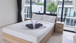 A modern hotel room with a large bed, a wooden headboard, a nightstand, and a view of buildings through large windows. - laguna 5 卧室，私人泳池别墅 (Tambon Choeng Thale)