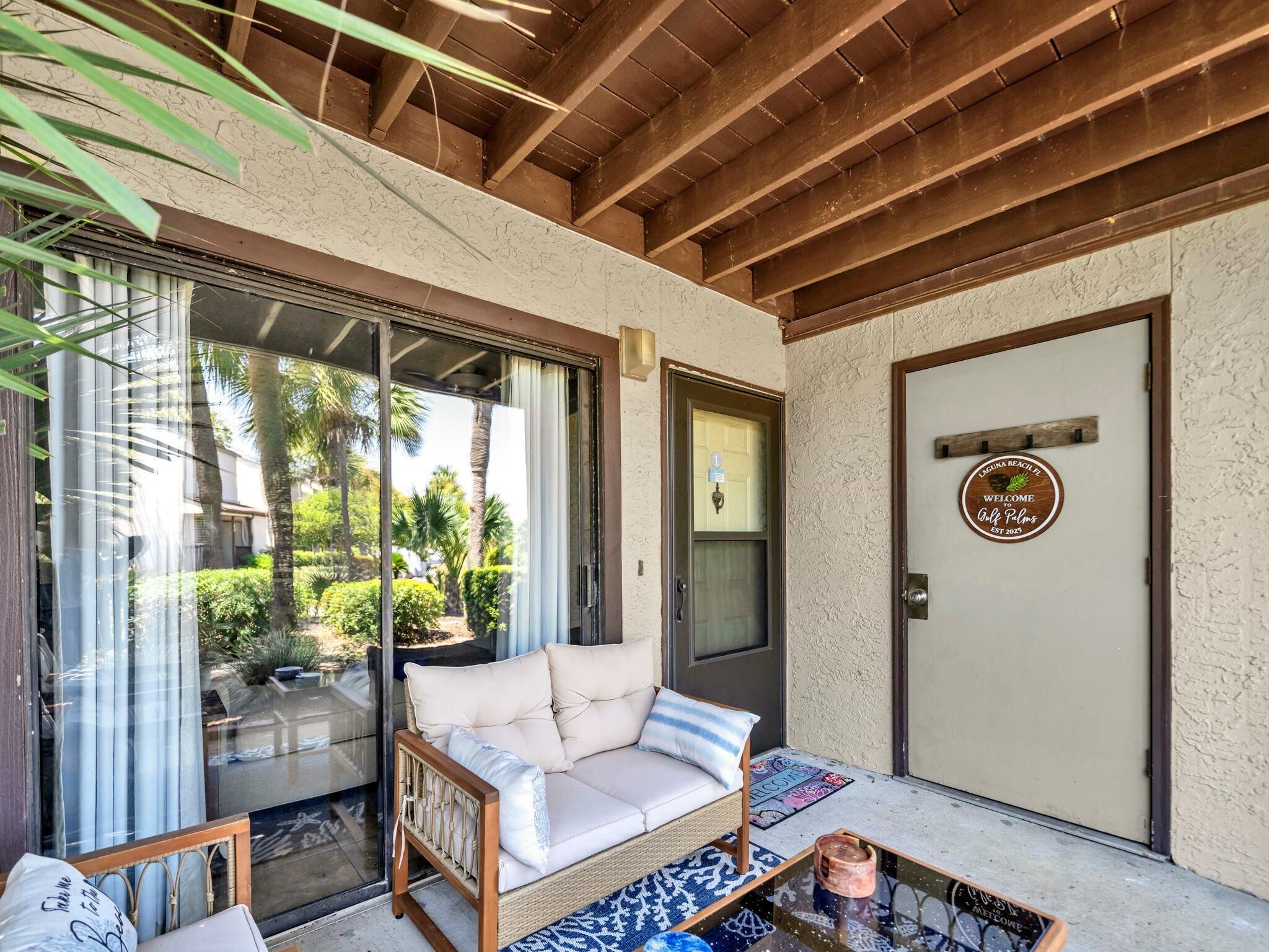 A cozy outdoor seating area with a cushioned sofa and a wicker chair, a glass door leading to a lush garden, and a wooden door with a welcome sign.