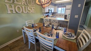 A dining area with a wooden table and white chairs, a kitchen in the background, and a wall with 'BEACH HOUSE' letters. - Portside I1- Gulf Palms (Panama City Beach)