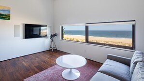 A modern living room with a large window overlooking a beach, a round white table, and a grey sofa. - Oceanview 5-BR House in Bethany Beach w/ Elevator (Bethany Beach)