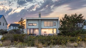 A modern two-story house with large windows and a flat roof, surrounded by natural vegetation and other houses. - Oceanview 5-BR House in Bethany Beach w/ Elevator (Bethany Beach)