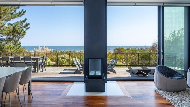 A modern interior with a large window overlooking the sea, a fireplace, and outdoor seating. - Oceanview 5-BR House in Bethany Beach w/ Elevator (Bethany Beach)