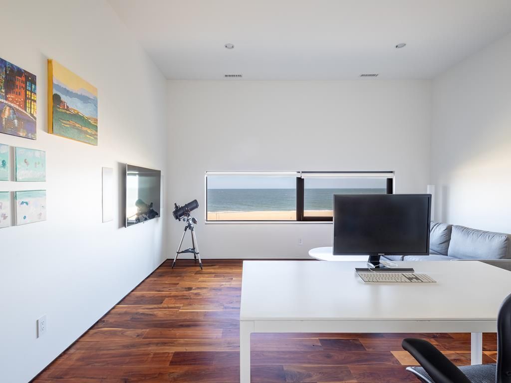 A modern office space with a large window overlooking a beach, a white desk with a computer, and a couch.