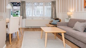 A modern living room with a sofa, a dining table, and a chair. There is a window with curtains, a radiator, and a lamp on a side table.