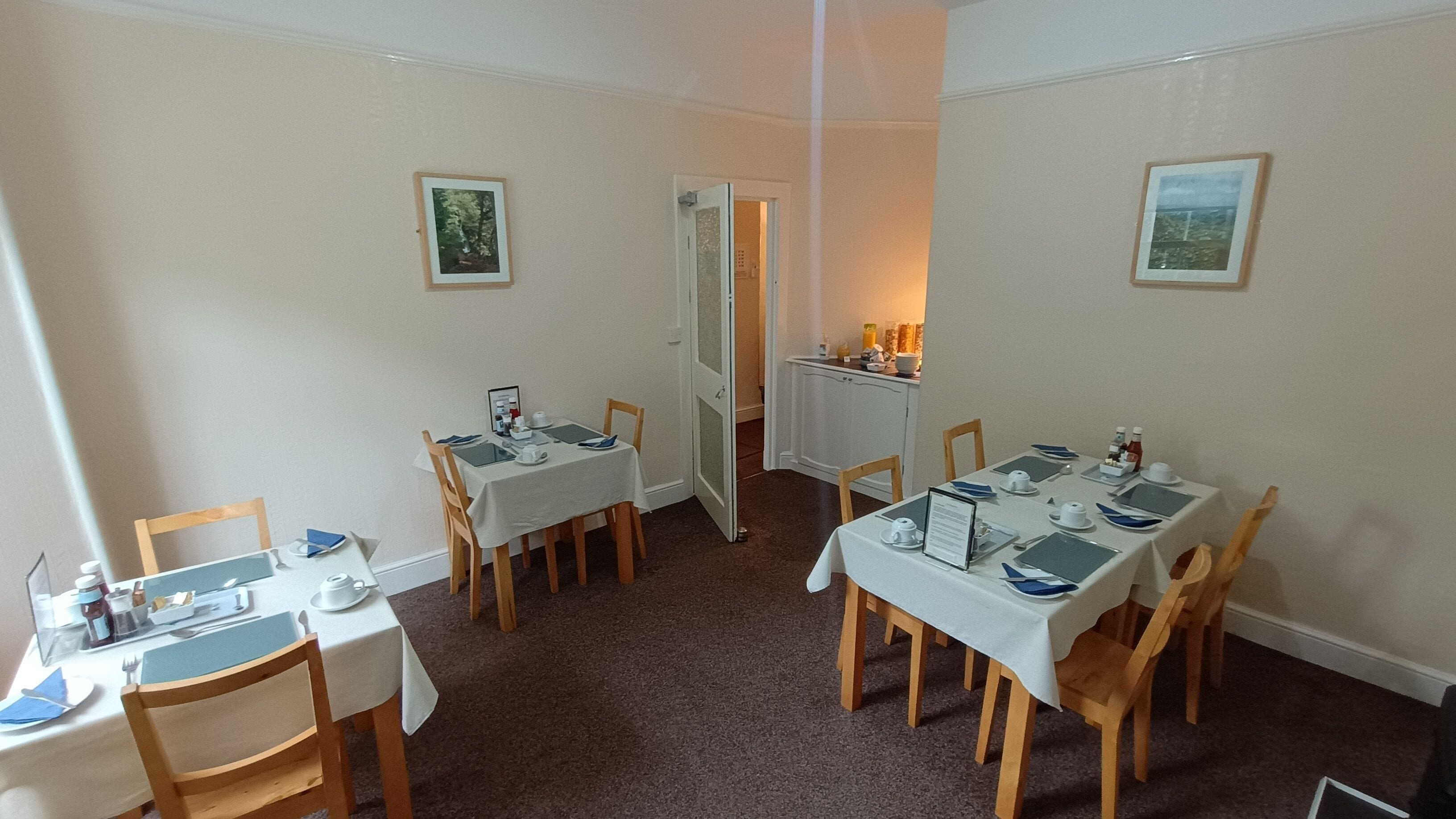 A dining area with four tables, each set with plates, cutlery, and glasses. There are wooden chairs around the tables. The room has white walls and a carpeted floor. Two framed pictures are hung on the walls.