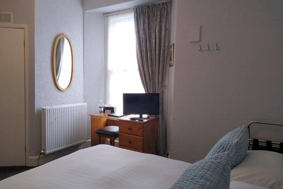 A hotel room with a bed, a dresser, a mirror, a window with curtains, a radiator, and a television.