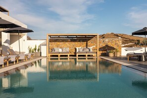 A pool area with lounge chairs and umbrellas, a stone wall, and a view of a hilly landscape. - Chora Kythnos Suites (Kithnos)
