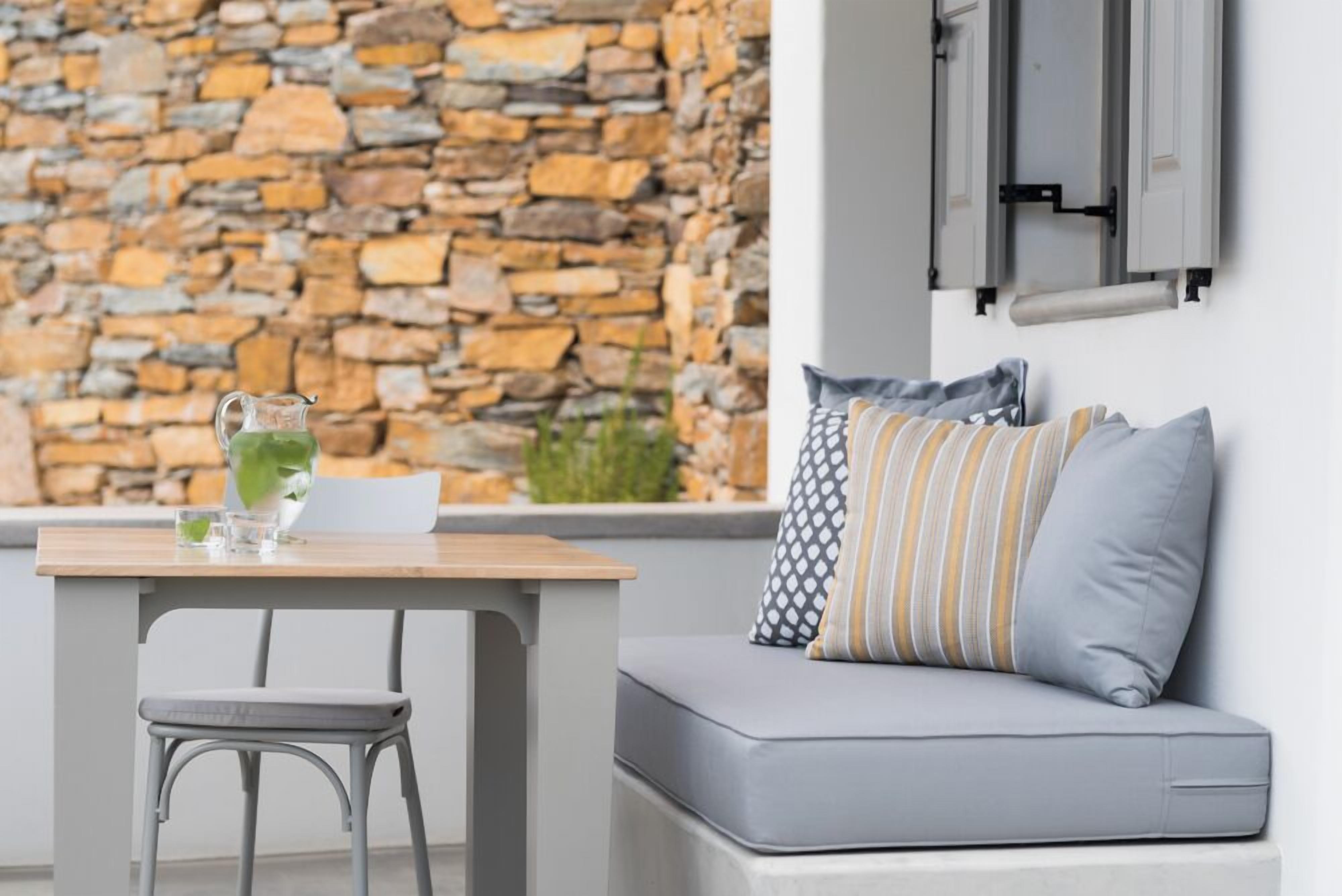 A modern room with a stone wall, a gray sofa with patterned cushions, a wooden table with a pitcher and glasses, and a metal chair.