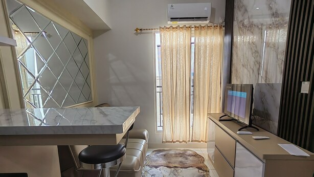 A modern kitchen with a marble countertop, a bar stool, a marble-patterned rug, and a television on a cabinet. - Podomoro Golf View By Arsakha Property (Gunung Putri)