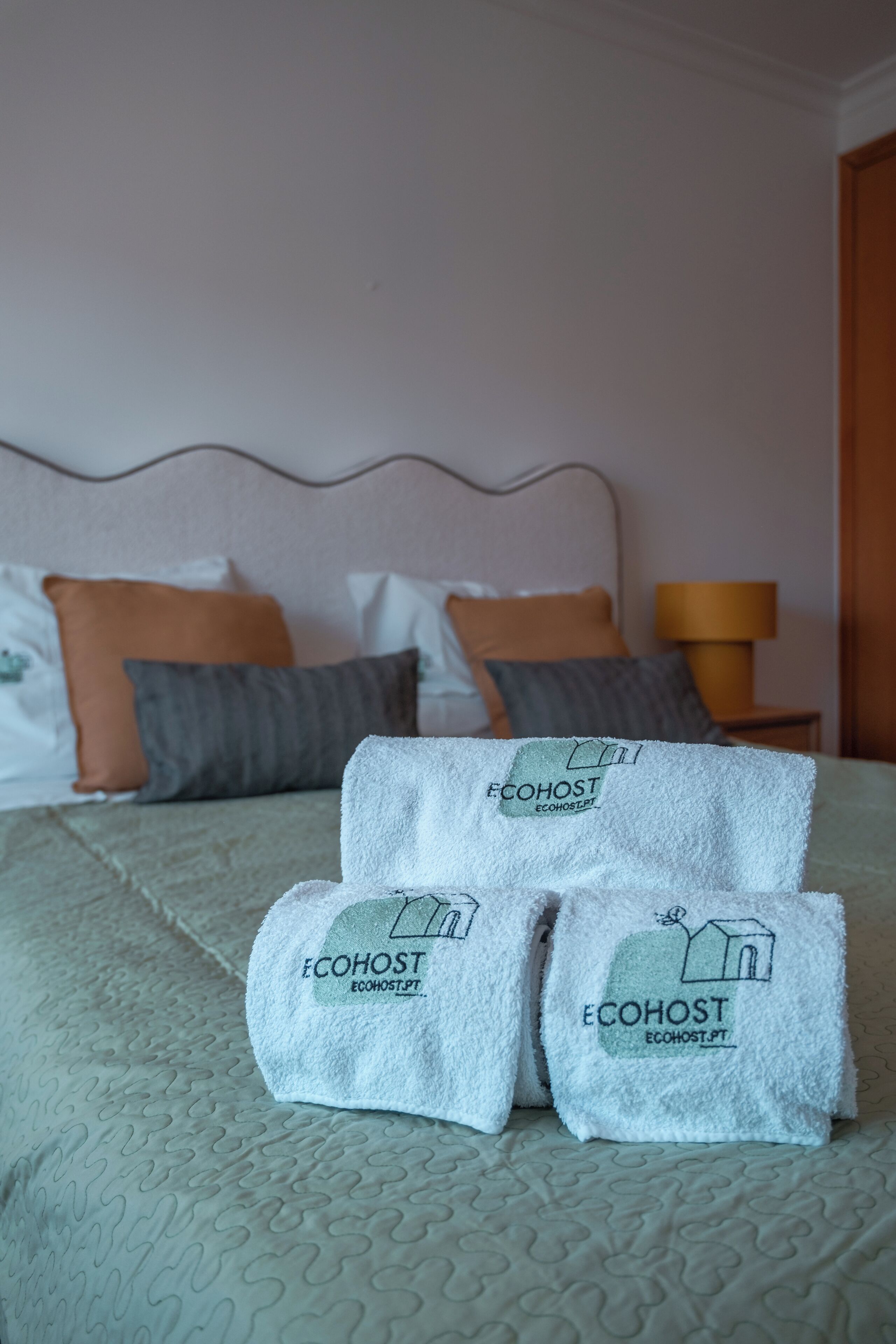 Una cama bien hecha con una colcha estampada, dos almohadas y tres toallas dobladas con la marca 'ECOHOST'.