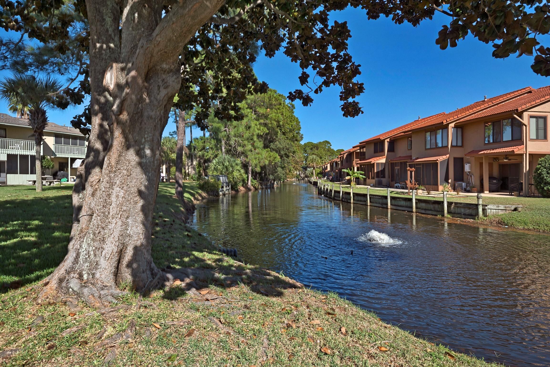 A river with a tree in the foreground, a wooden bridge, and a row of buildings with a red roof along the riverbank.