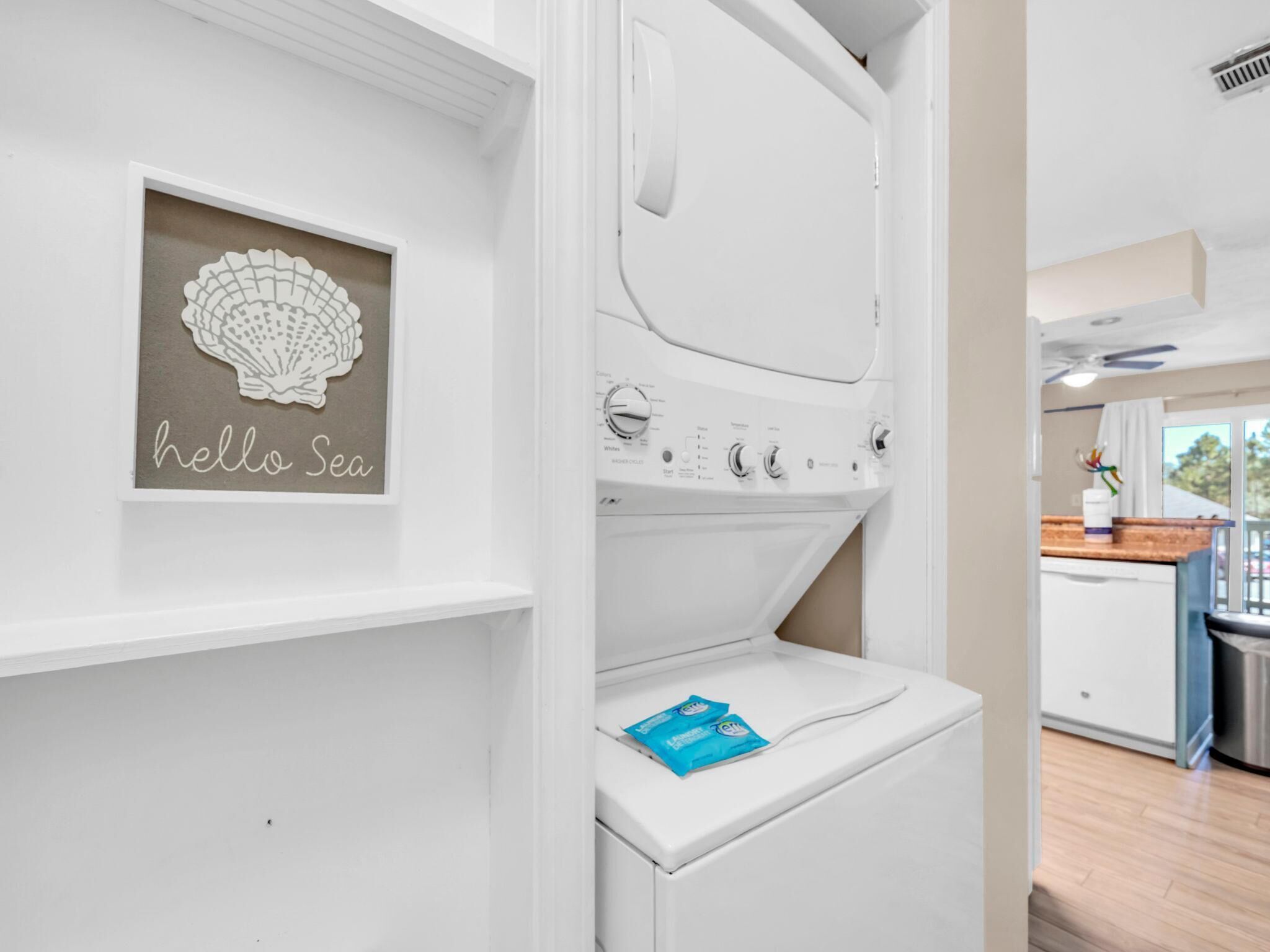 Une buanderie avec une machine à laver et un sèche-linge fixés au mur, une œuvre d’art encadrée dont le cadre est décoré d’un motif festonné, et un sachet de lessive bleue posé sur le sèche-linge.