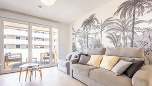 Una sala de estar con un sofá, una mesa de centro y una silla. Se aprecia una ventana grande con vistas a un edificio.