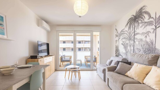 Una sala moderna con sofá, mesa de comedor y televisor. Posee un ventanal corredero que da a un balcón con mobiliario exterior.