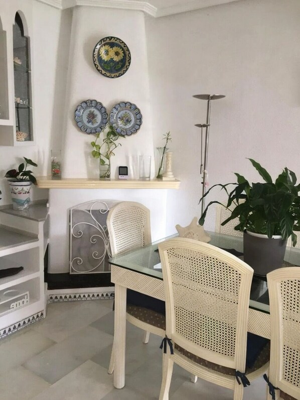 A dining area with a glass table and wicker chairs, a shelf with decorative plates, and a potted plant.