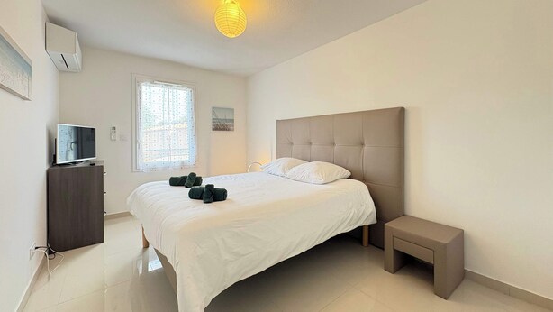 Un dormitorio con cama, mesita de noche, televisor sobre un mueble y ventana con cortinas.