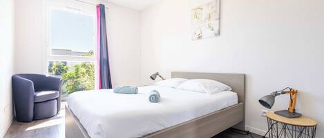 Ein modernes Schlafzimmer mit einem Bett, einem Schreibtisch mit Lampe, einem Sessel und einem Fenster mit Vorhängen.