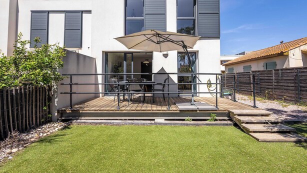 Une maison moderne à deux étages, dotée d’une terrasse, de mobilier d’extérieur et d’un grand parasol.