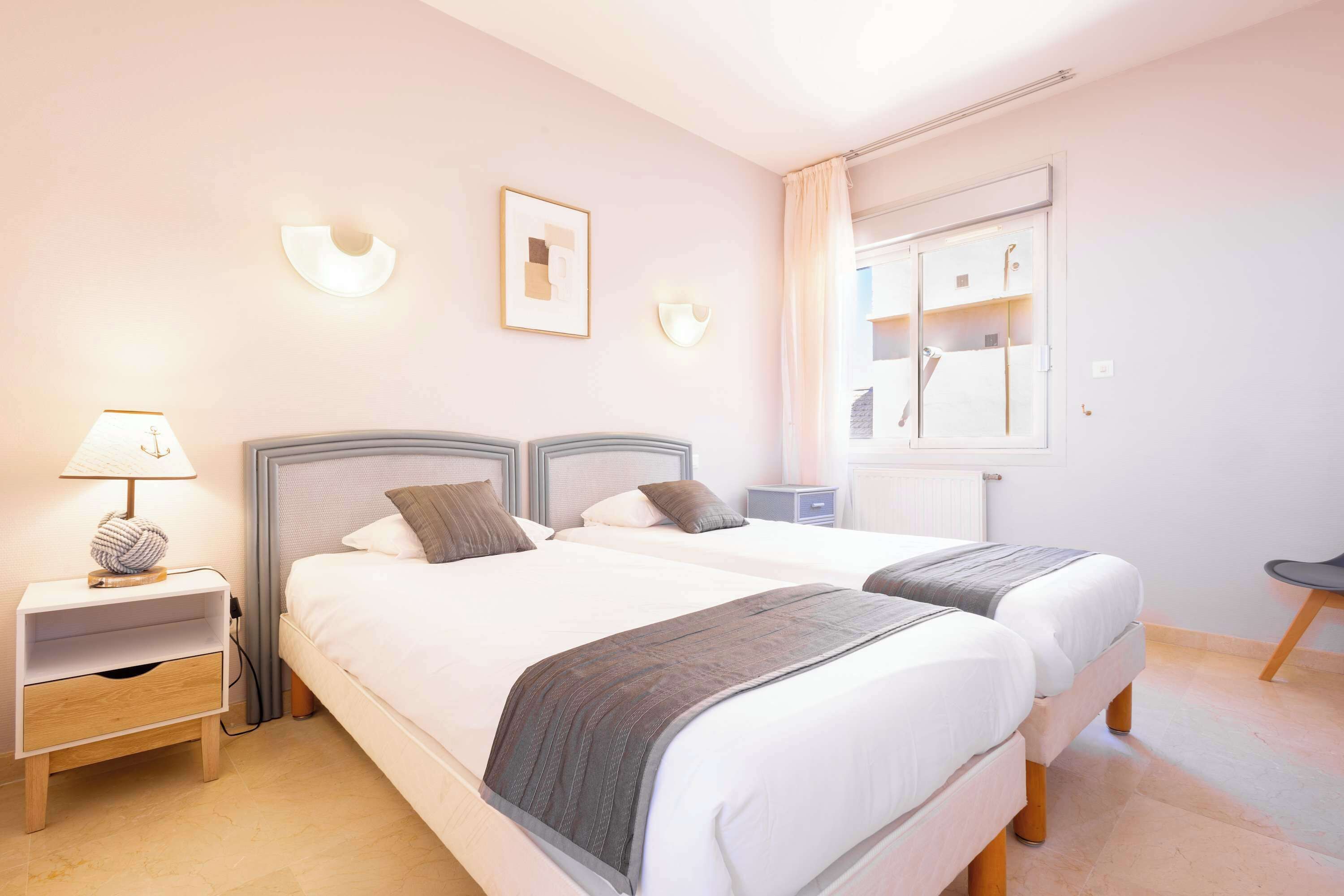 Un lit double avec une tête de lit grise, une table de chevet avec une lampe et une petite table de nuit avec un tiroir.