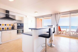 A modern kitchen with a central island, bar stools, and a view of the sea through large windows.