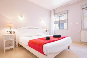 A bedroom with a white bed, red blanket, two bedside lamps, and a window with blinds.