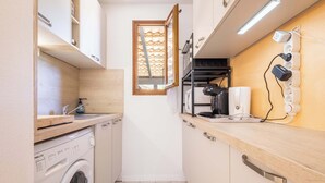 A compact kitchen with a washing machine, a built-in oven, and a window with a wooden blind.