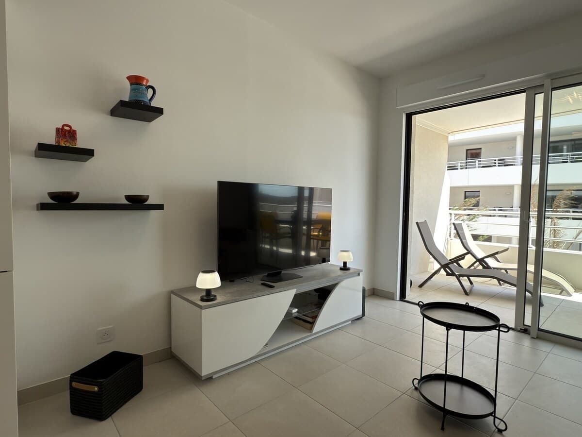 A modern living room with a flat-screen TV, a white console table, and a small round table. There are shelves with decorative items and a balcony with chairs.