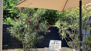Ein Patio mit einer weißen Lounge-Liege unter einem großen, beige farbenen Sonnenschirm, umgeben von Pflanzen und einer schwarzen Wand.