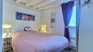 Ein Schlafzimmer mit Bett, Nachttisch, Lampe, Wandregal und einem Fenster mit Vorhängen.