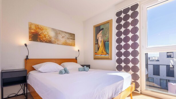 A bedroom with a wooden bed, white bedding, a nightstand, a wall-mounted painting, and a window with a view. - Villa Bianca – Spacious Apartment (Sérignan)
