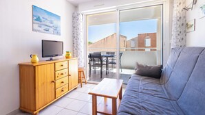 A living room with a sofa, a wooden cabinet, a television, and a sliding glass door leading to an outdoor patio. - Le Clos Marin – Beautiful Flat in Secure Residence (Valras-Plage)