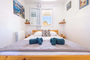 A neatly made bed with two towels, a window with a view, a painting on the wall, and a shelf with items. - Le Clos Marin – Beautiful Flat in Secure Residence (Valras-Plage)