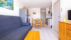 A compact living space with a sofa, dining table, chairs, a wooden cabinet, a TV, and a kitchen area. - Le Clos Marin – Beautiful Flat in Secure Residence (Valras-Plage)