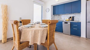 A dining area with a round table set for four, surrounded by wicker chairs. A kitchen with blue cabinets and stainless steel appliances is visible in the background. - Valras 'Loc Archipel Panarea - Cap Horizon (Valras-Plage)