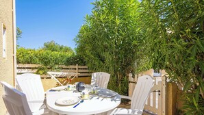 A round table with white chairs, a teapot, cups, and plates, set outdoors with a wooden fence and greenery in the background.