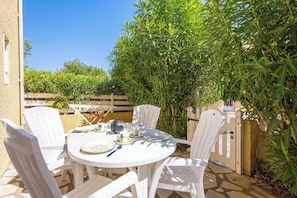 A round table with white chairs, a teapot, cups, and plates, set outdoors with a wooden fence and greenery in the background.