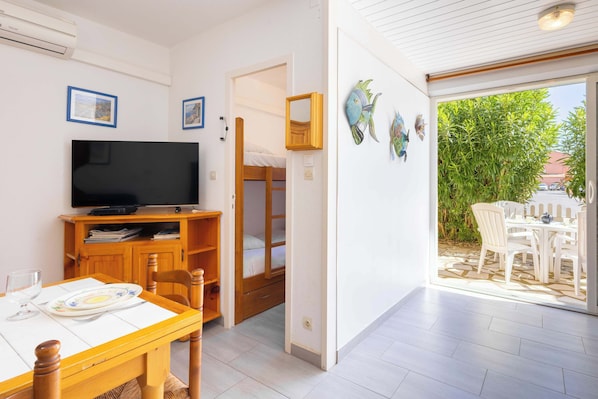 A room with a TV, wooden furniture, a dining table, and a view of an outdoor patio with chairs.