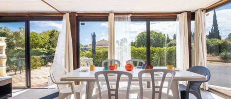 A dining area with a table and chairs, large windows with curtains, and a view of greenery and buildings outside.