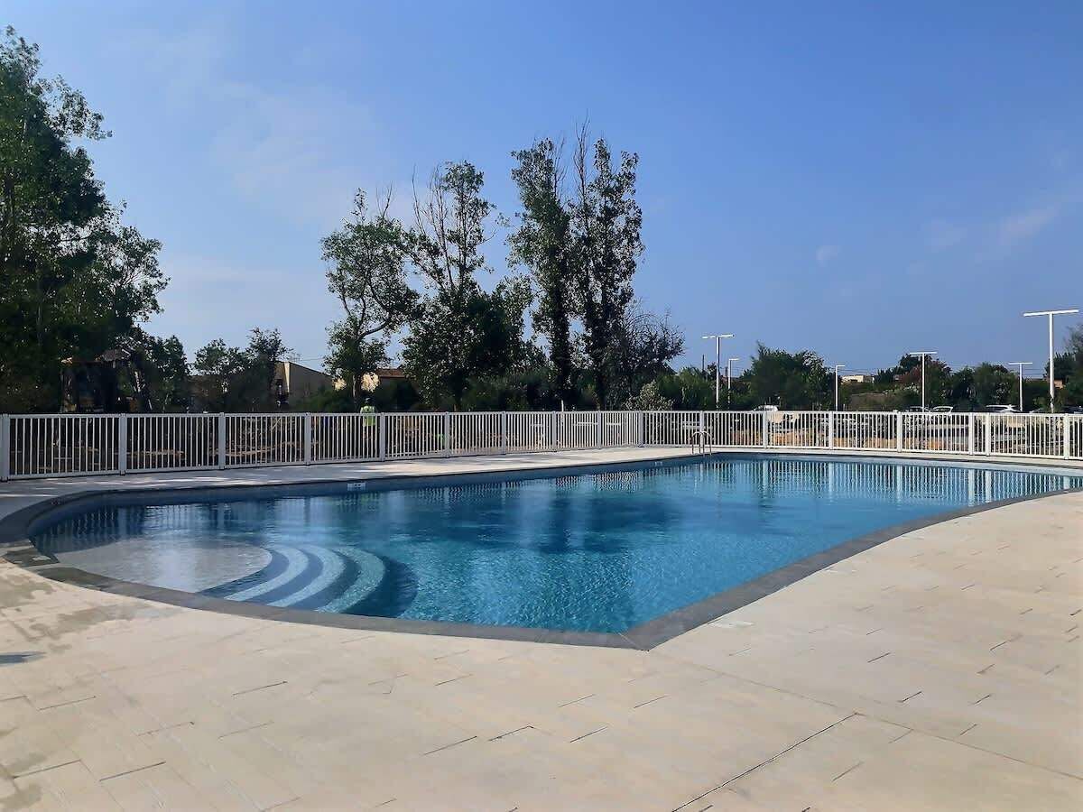 A rectangular swimming pool with a white fence surrounding it, set on a paved area with trees in the background.