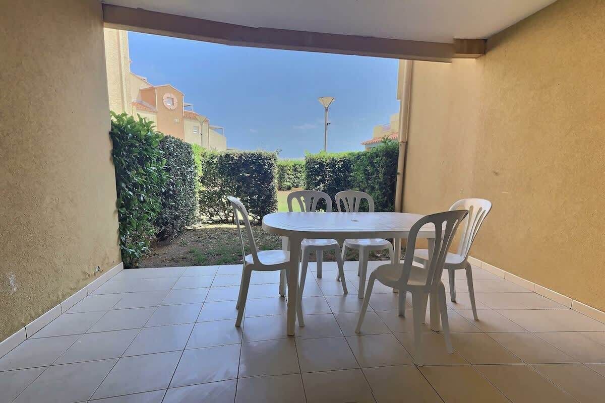 A patio with a white table and chairs, a tiled floor, and a view of a garden and buildings in the background.