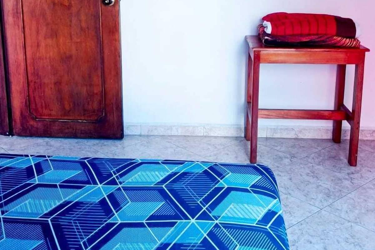 Une pièce avec une porte en bois, un lit recouvert d’une courtepointe à motifs géométriques, une petite table en bois avec un coussin rouge, et un téléviseur fixé au mur.