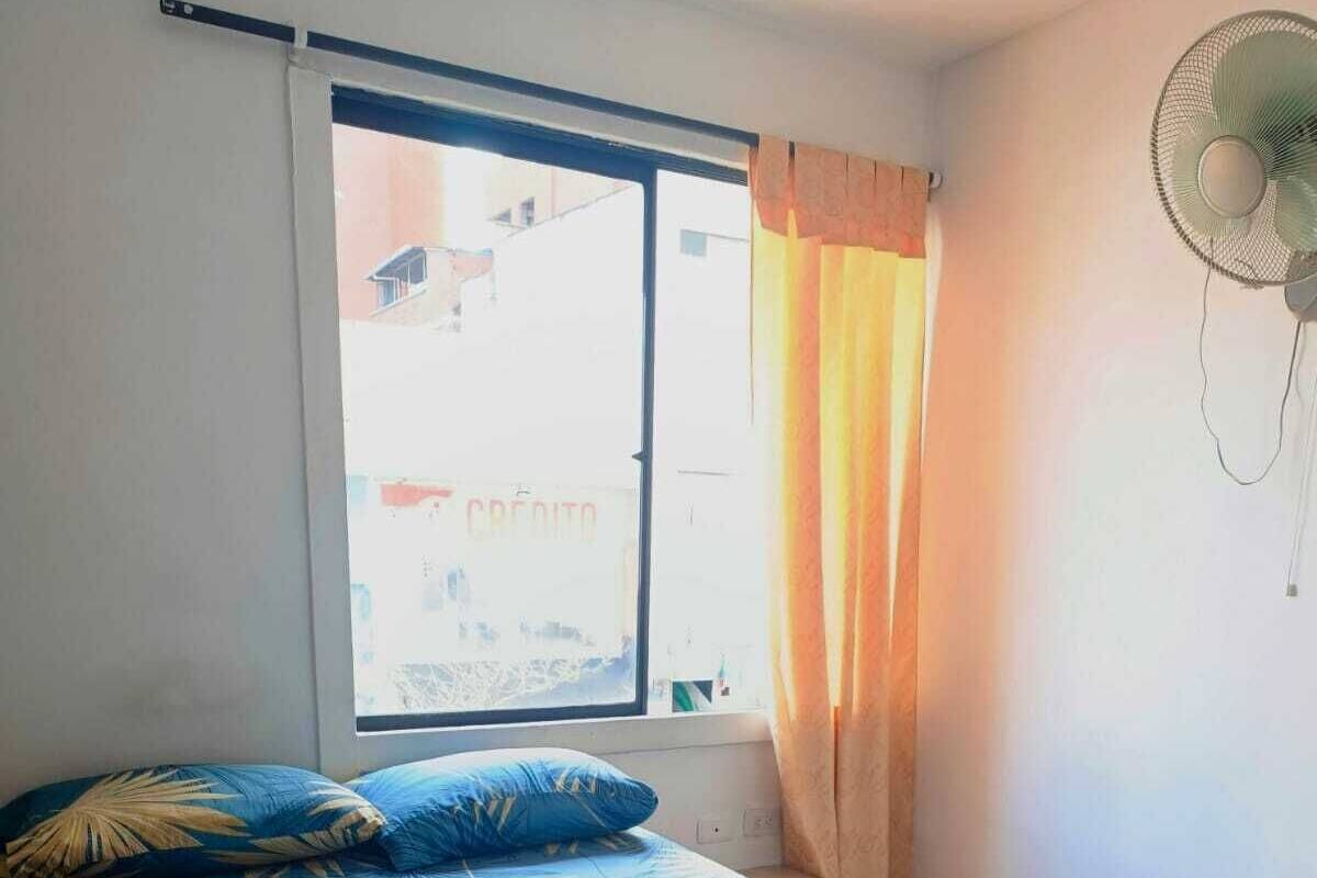 Une chambre à coucher avec un lit, une fenêtre avec des rideaux et un ventilateur fixé au mur.