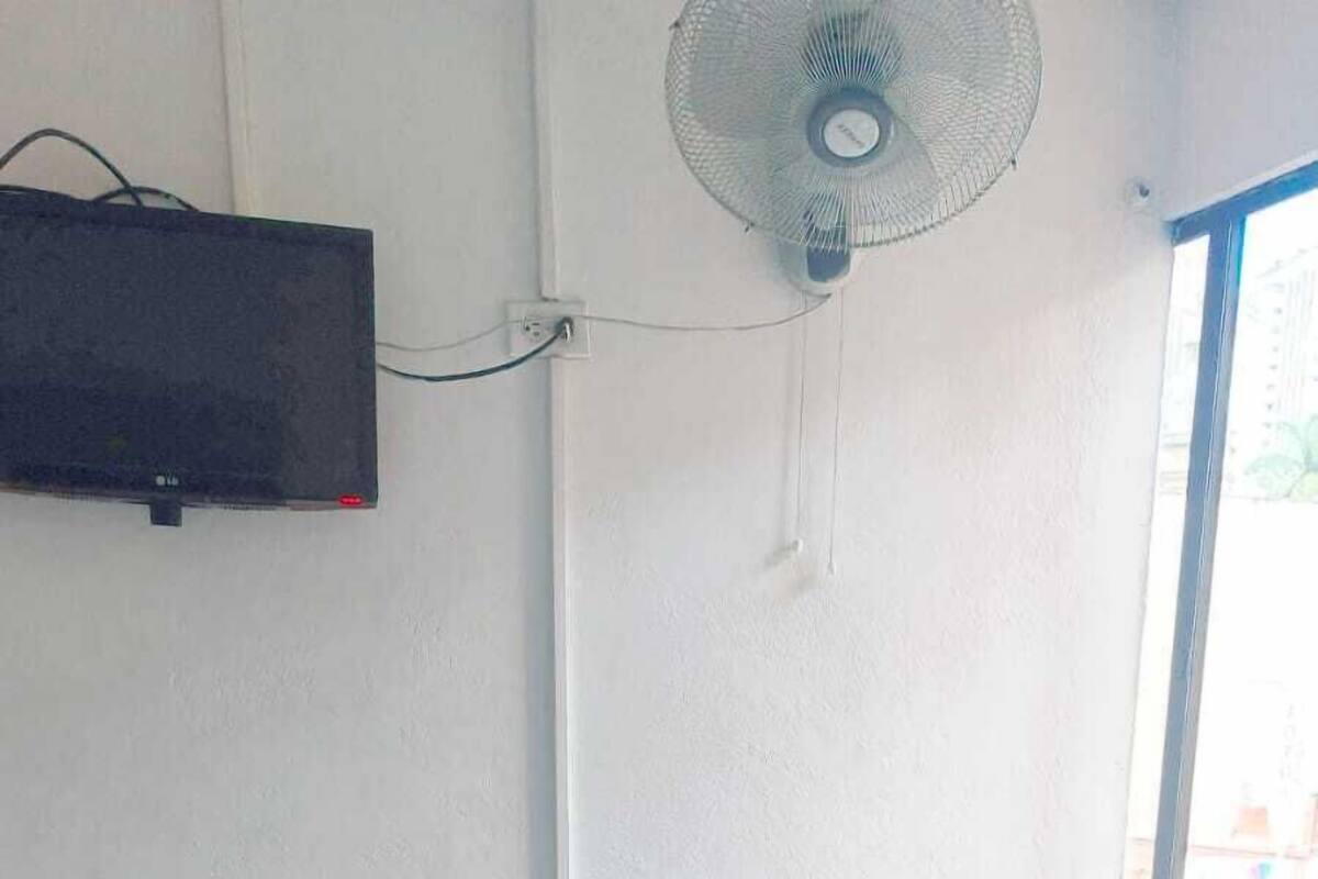 Une chambre avec un lit, un téléviseur fixé au mur, un ventilateur sur pied et une fenêtre.