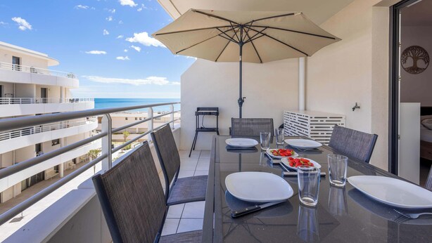 Un balcón con una mesa lista para dos personas, vista a la playa y un paraguas grande.