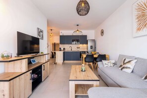 A modern living room with a flat-screen TV, wooden furniture, a grey sofa, and a kitchen in the background. - Valras'Loc Archipel Panarea Sea View and Comfort (Valras-Plage)